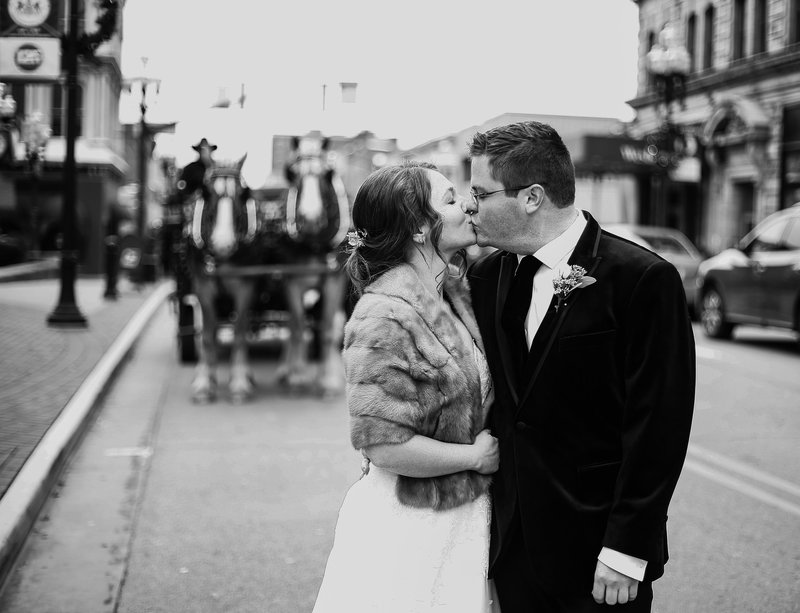 Bride and groom kiss in the street with horse drawn carriage in the background