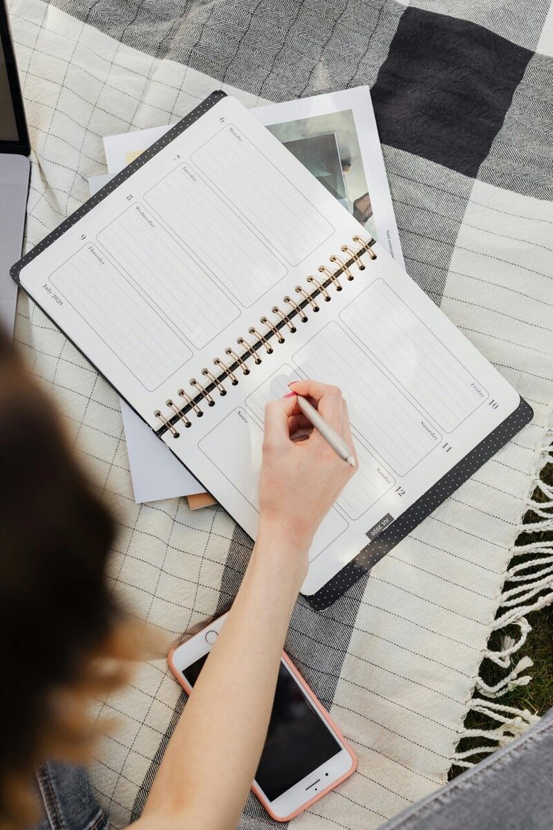 Person writing in a weekly planner while sitting on a picnic blanket, with a phone and notebook nearby.