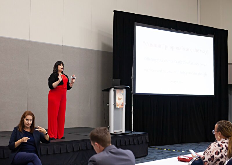 Speaker, Renee Dalo, presents on stage at a conference in red jumpsuit and black cardigan