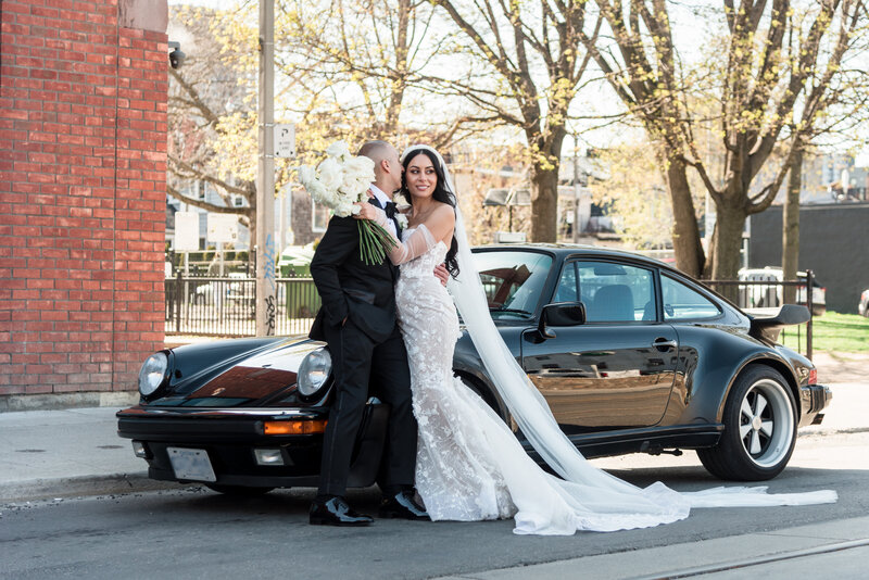 The bride and groom are leaned over the side of this black classic Porsche as the groom is about to kiss her cheek, white roses and flowers on the brides bouquet and an elegant and modern dress