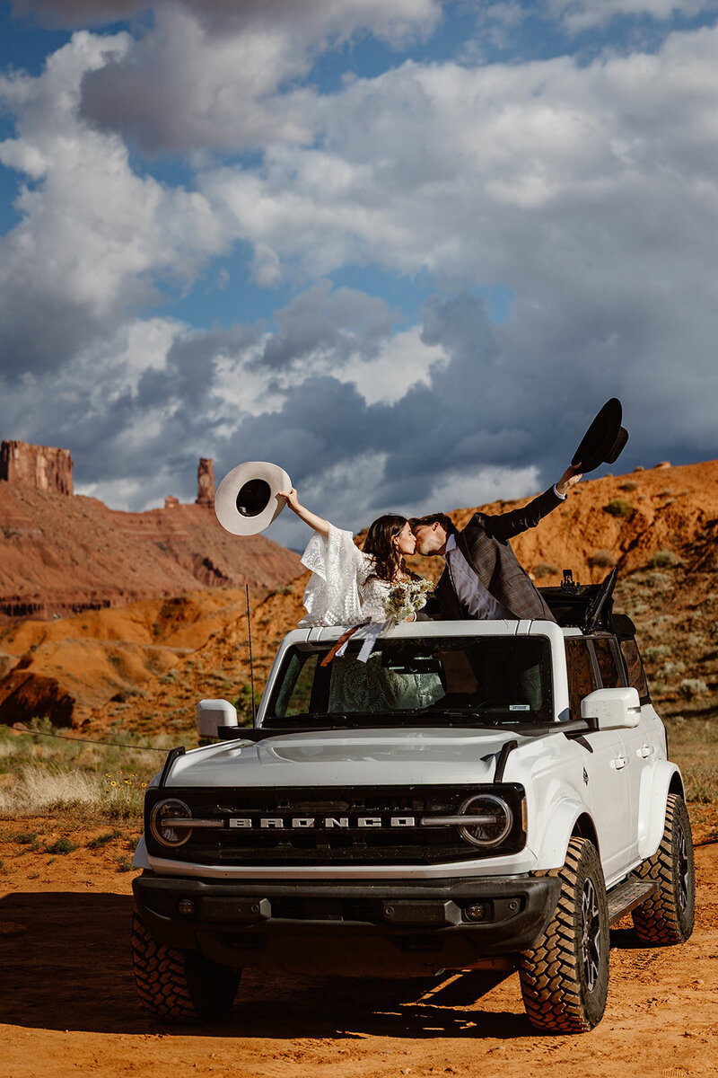 A couple in the Bronco at Ida Gulch in Moab. They are standing out of the sunroof, kissing, and holding their hats up