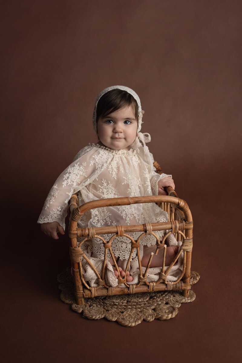 A young toddler sits in a wicker crib in a studio wearing a bonnet and lace dress taken by a Lafayette Baby Milestone Photographer