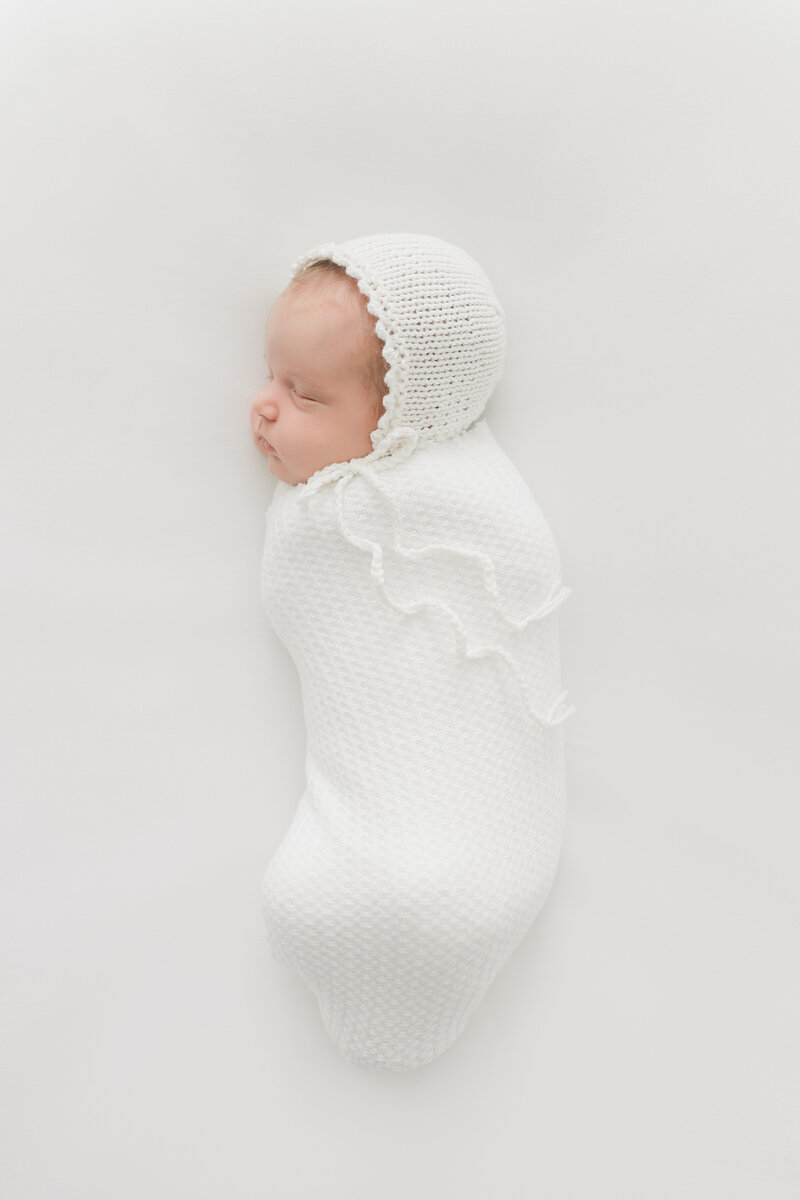 A beautiful baby girl swaddled in white on a white backdrop with a white bonnet on her head by northern va family photographer
