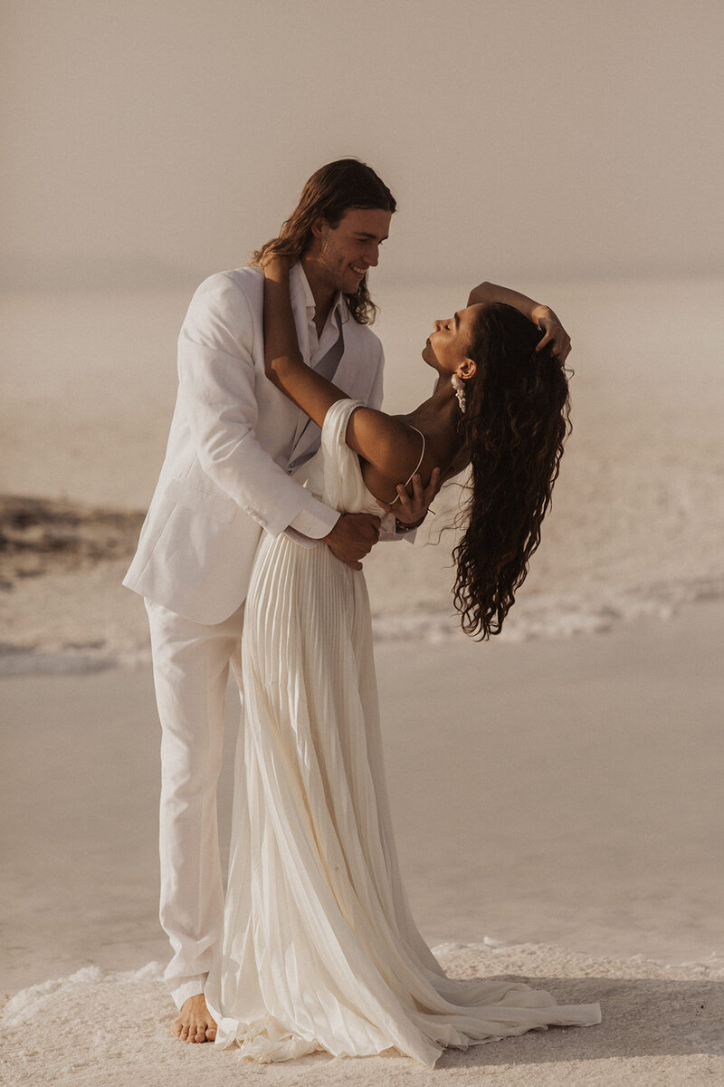 A couple in wedding attire intimately interacting on a sandy beach.