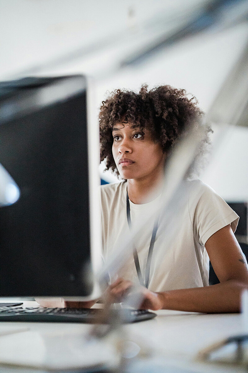 A person with curly hair focuses intently on a computer screen in an office setting, conveying concentration and productivity. Blurred foreground elements add depth.