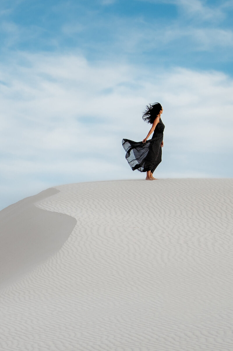 A woman from San Diego standing in a black dress on top of the sand dunes in New Mexico