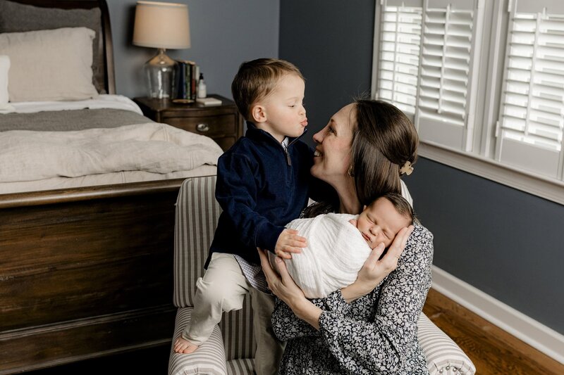 Little boy dressed in blue gives his mother a kiss during their lifestyle newborn session while Mom holds their newest little baby wrapped in white