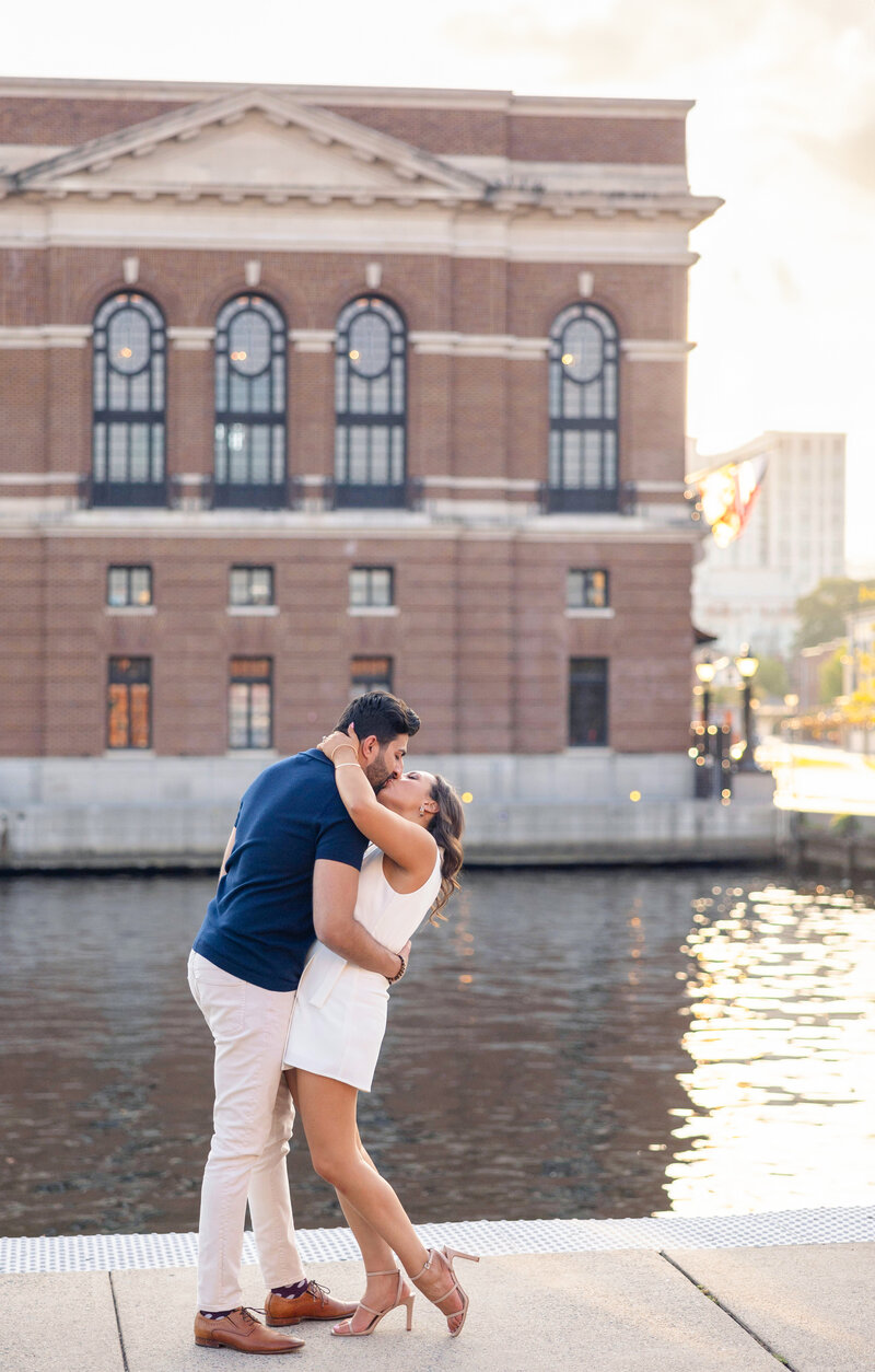 A couple embraces passionately by a waterfront, with a historic brick building in the background. The man wears a navy shirt and beige pants, while the woman is in a white dress with heels. The scene is lit by a warm, soft glow from the setting sun.