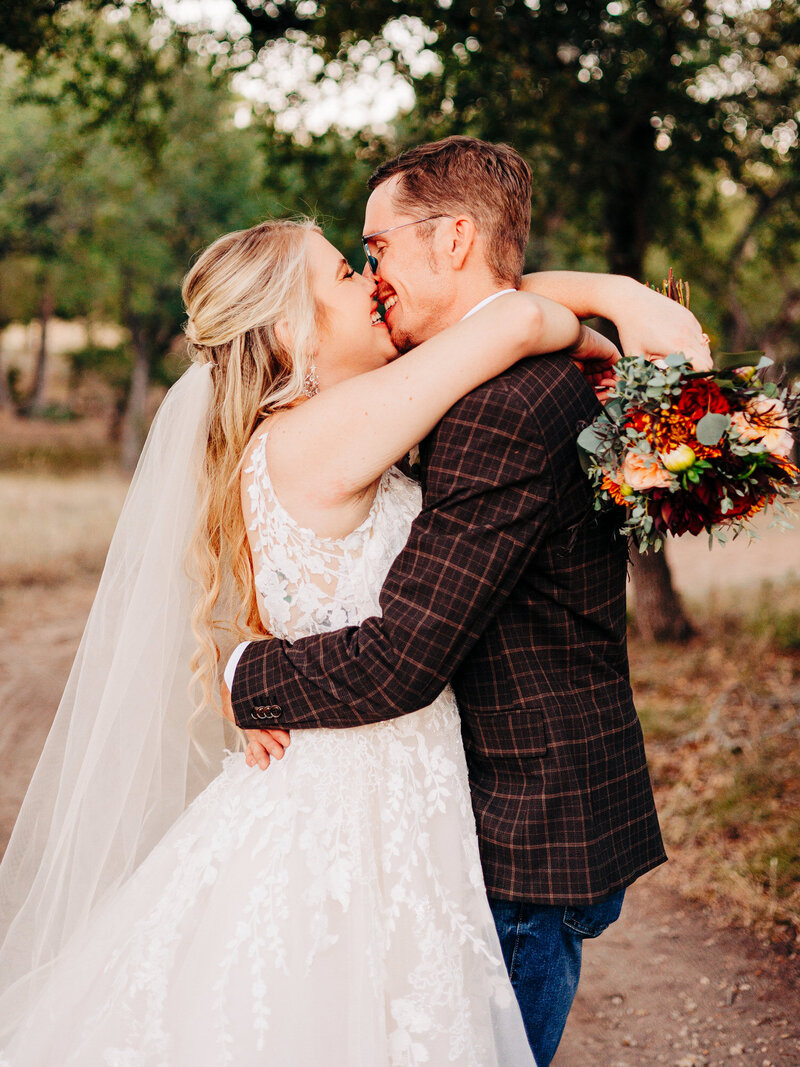 This image, taken by wedding photographer in San Antonio KD Captures, features a bride and a groom holding each other in a tight embrace. The woman is wearing a veil and has blonde hair. The man is wearing a brown plaid suit jacket. The woman holds a colorful bouquet as she wraps her arms around her groom's neck. The couple is just about to kiss while smiling at eachother.