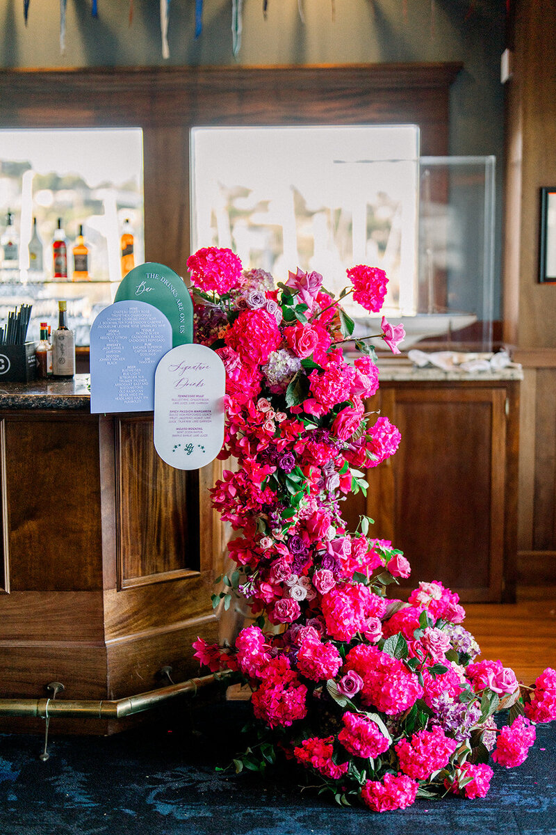Pink and red floral escort card installation spilling over bar at Tiburon Yacht Club wedding in California captured in film photography style