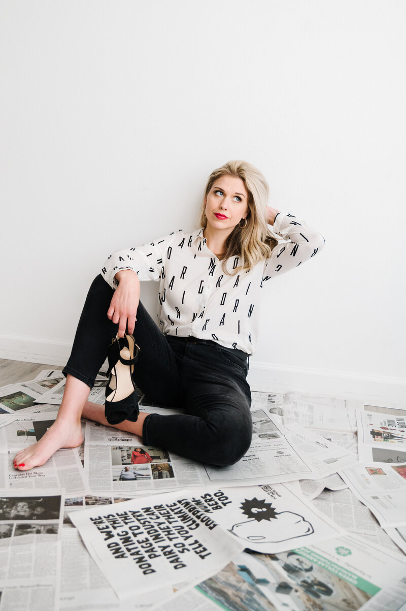 Sarah Klongerbo sitting against a white wall on a floor covered with newspaper pages