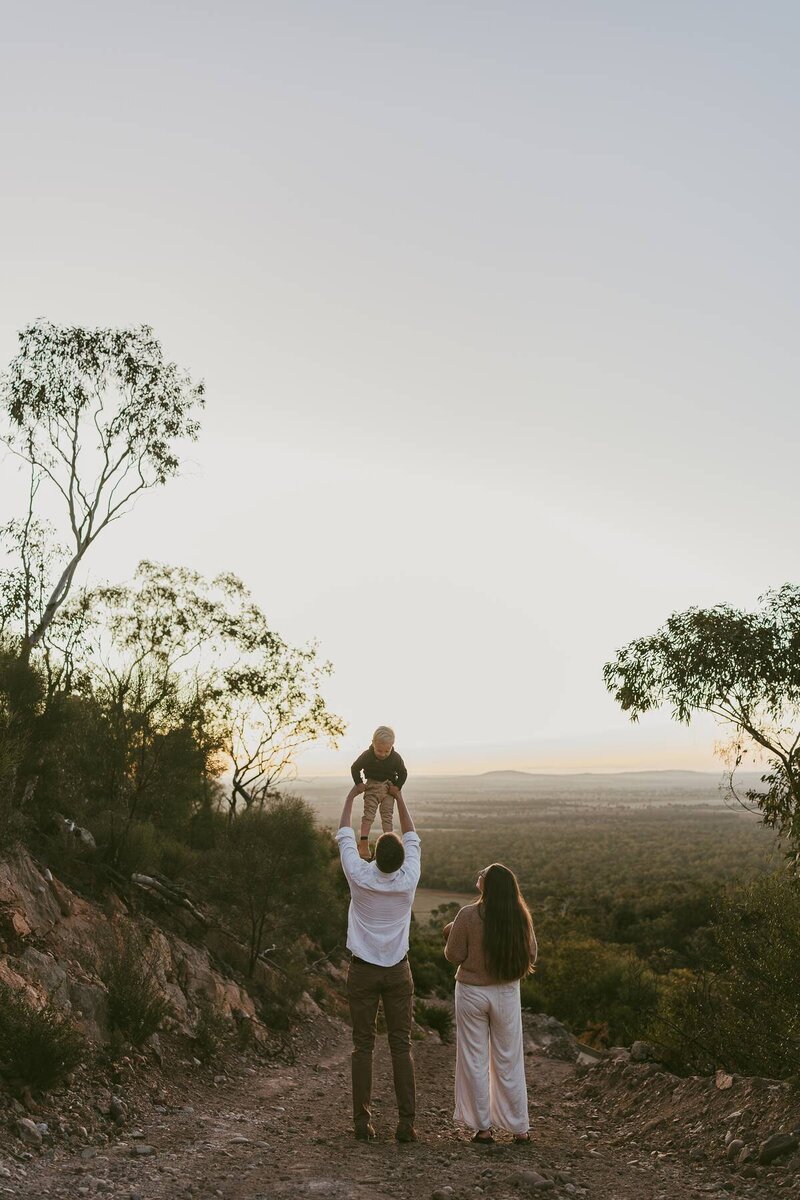 Family standing on a hilltop at sunset during a lifestyle family maternity session, with the father lifting a toddler into the air while the mother watches