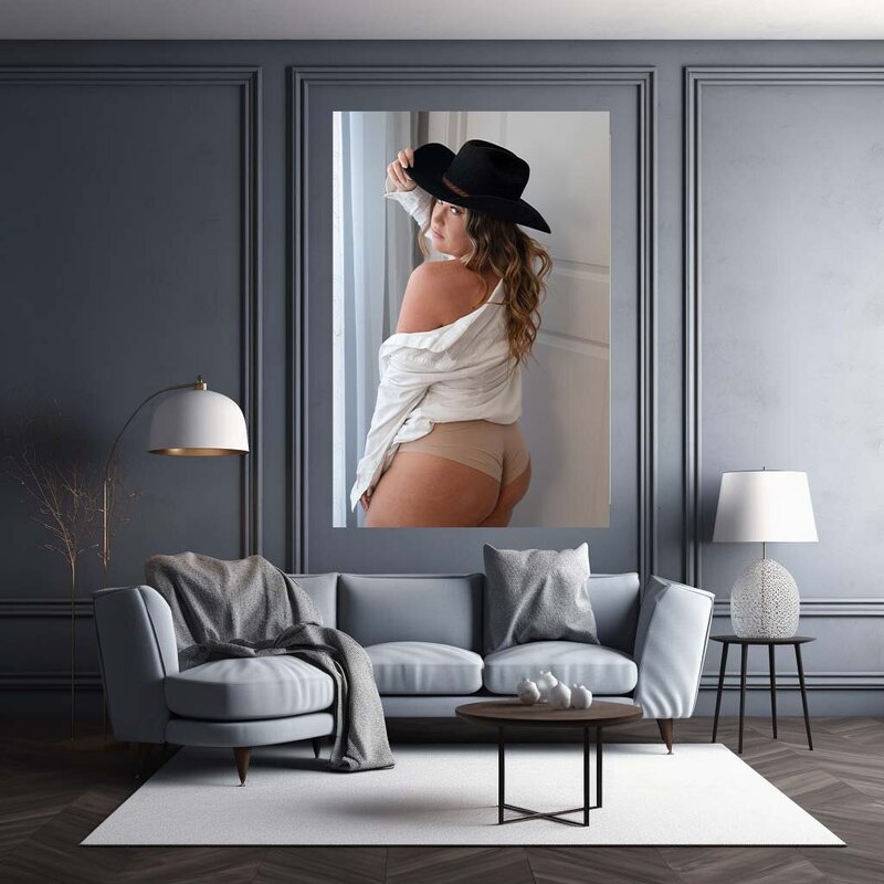 A canvas hanging on the wall of a woman wearing a white shirt that is off the shoulder and wearing a black hat and looking over her shoulder at the camera.