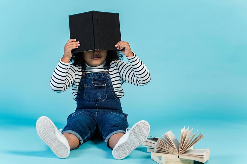 A child sitting on the floor with a light blue background, playfully holding a book over their face, wearing denim overalls and a striped shirt, with an open book lying next to them.