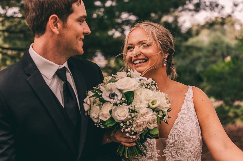 A bride and groom smiling at each other, holding a bouquet of white and pale blue flowers, in an outdoor setting orchestrated by a wedding coordinator in Iowa.