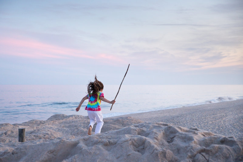 Young boy with long hair holds a stick and jumps off of the dunes at East Hampton Main Beach in East Hampton, NY.