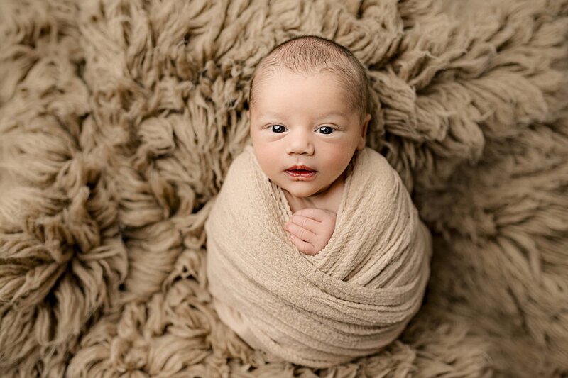 Baby has his eyes open as he looks up while wrapped in a brown wrap laying on a brown rug posed during his newborn photo session