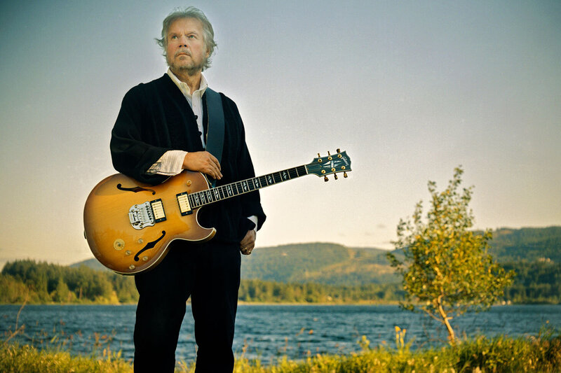 Randy Bachman musician portrait standing in front of lake arm resting on guitar slung over shoulder small tree beside him