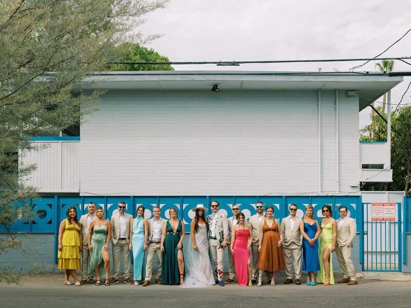 large wedding party in bright multicolored attire standing in front of a building with a blue patterned metal fence
