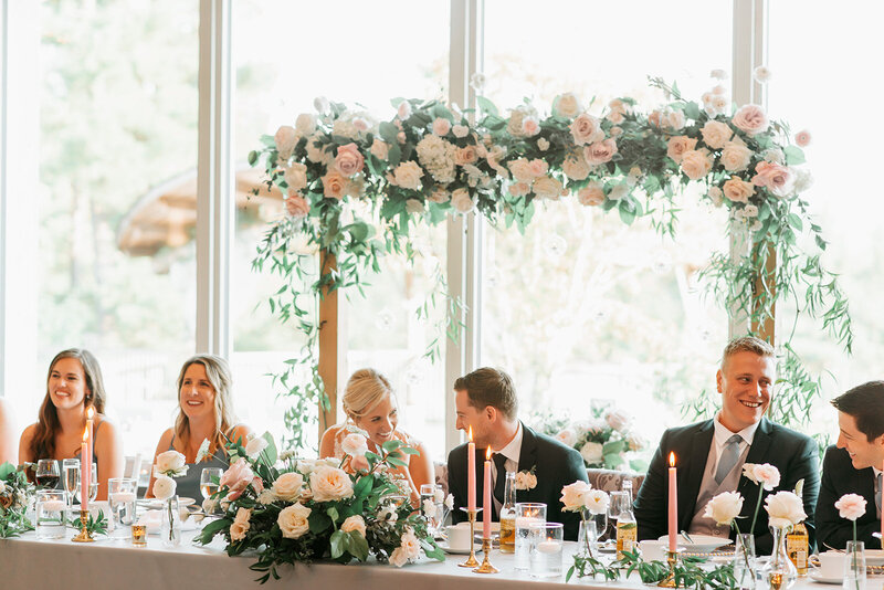 Newlyweds and their wedding party sit at a long head table with a large wood arch behind them covered in garden-inspired florals at a wedding at Le Belvedere venue in Wakefield Quebec