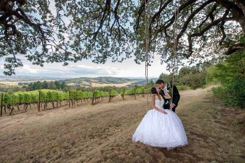 a groom leans down to kiss a bride on a swing overlooking vineyards at youngberg hill