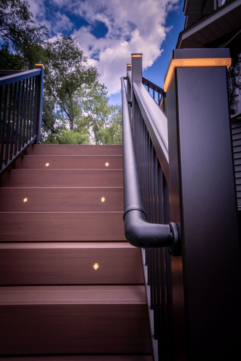 PVC deck staircase with riser lights on alternating steps, black aluminum picket railing with post cap lights, and an ADA rail. 