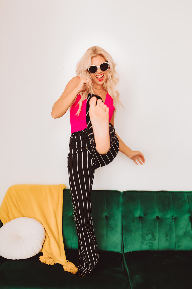 Founder of Infinite Productions, Andi Sweeny, standing on a green couch wearing black and white striped pants, a bright pink shirt, and sunglasses. She is kicking one foot into the camera.