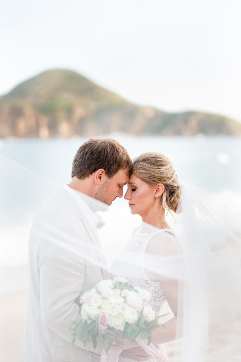 A bride and groom sharing a tender moment on the beach with their foreheads touching, the bride holding a bouquet, captured by a luxury wedding photographer.