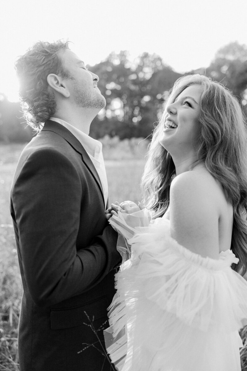 A joyful couple laughing together in a field, with the man looking upwards and the woman looking at the man, both in formal attire, captured by a luxury wedding photographer.