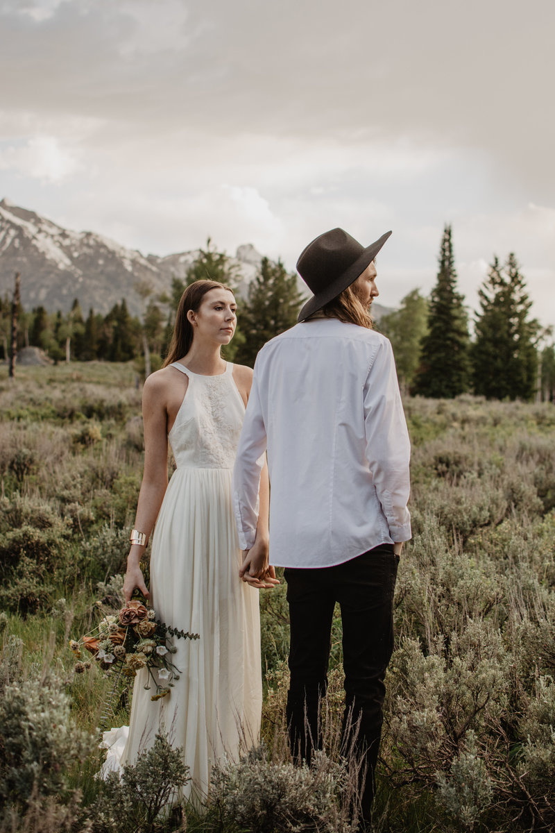 wyoming elopement with boho bride and trendy groom stand in a field right outside of the Tetons holing hands. Groom is wearing a black hat and a light blue button down and is looking in the distance. Bride is holding her wedding bouquet and looking at her groom