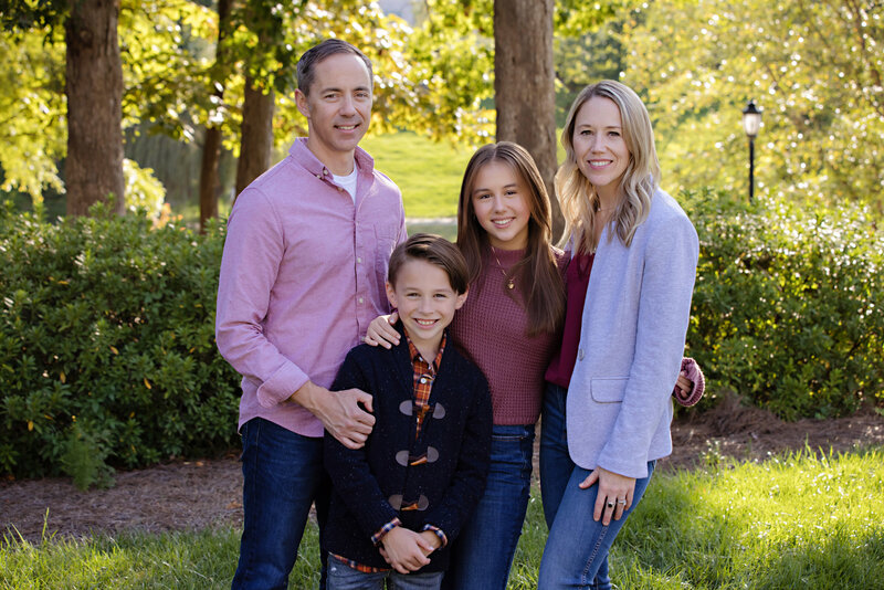 Lovely photograph of a family of four looking at the camera smiling while standing in a park setting with greenery and trees behind them.