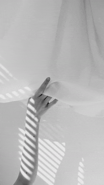 A moving image of a woman's hand  delicately holding some white fabric in reflected light through a blind which creates striped shadows.