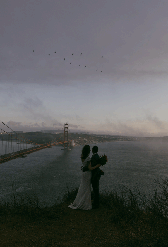 bride and groom standing on cliff above Pacific ocean with golden gate bride in the background