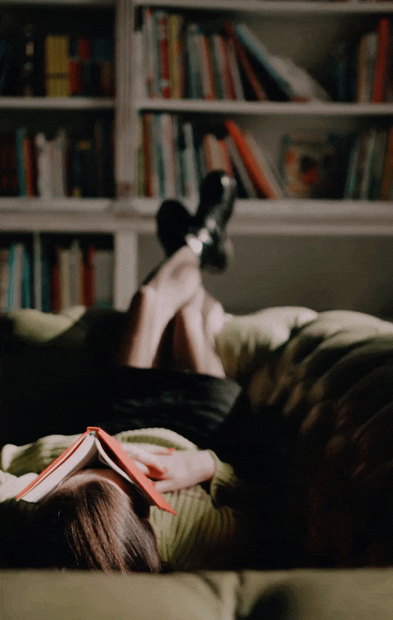 A woman laying on a green couch with a book covering her face in a library