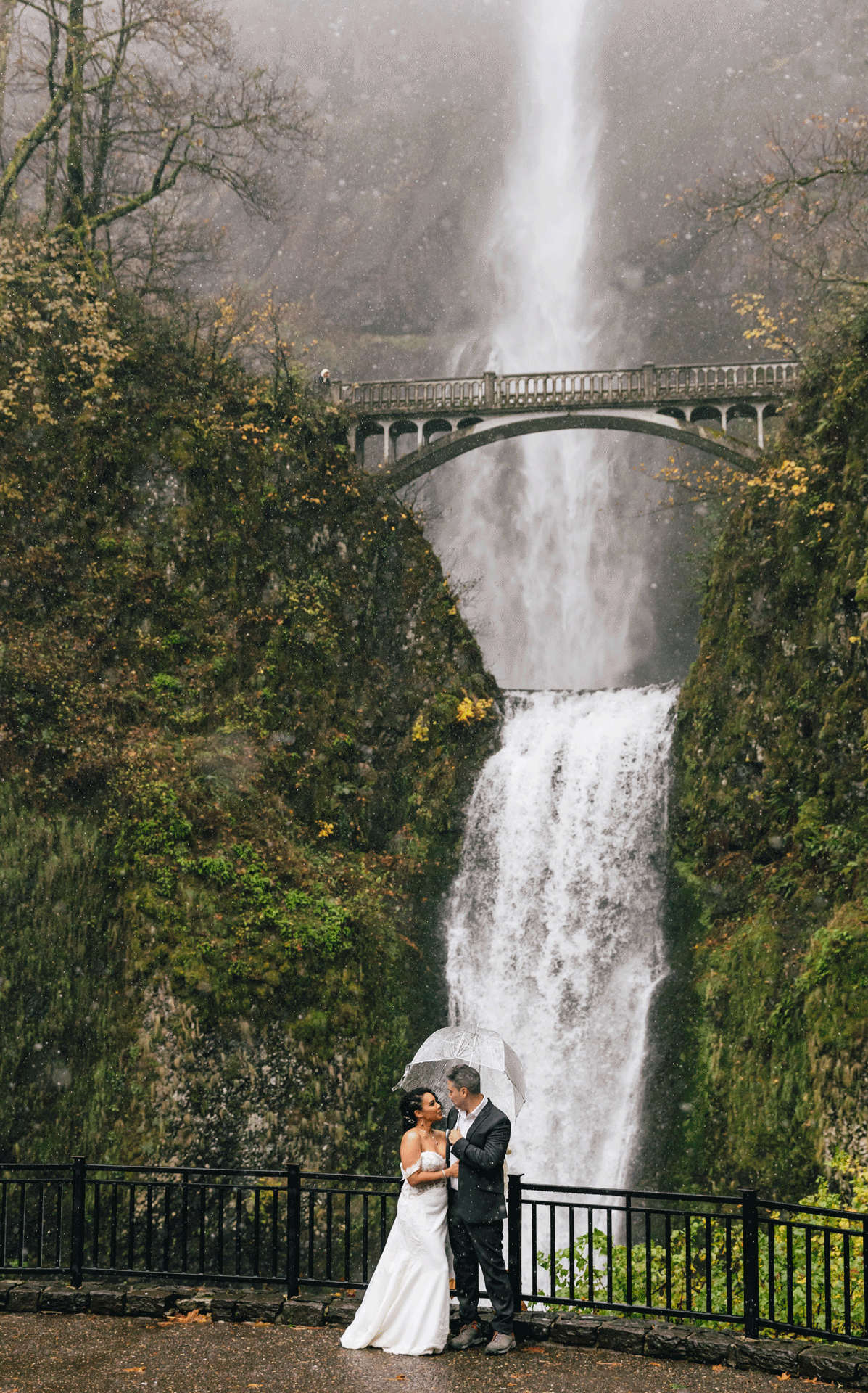 A Puerto Rican bride wearing a white gown with the sleeves off shoulder stands under a clear umbrella held by her groom in a black suit standing in front of Oregon's Multnomah Falls as the snow begins to fall for their winter adventure elopement. | Erica Swantek Photography