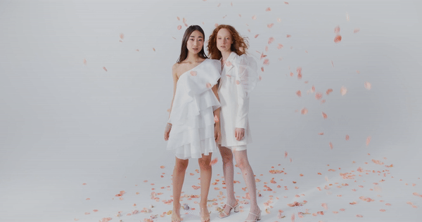 two women in white dresses look at the camera as pink petals fall to the ground