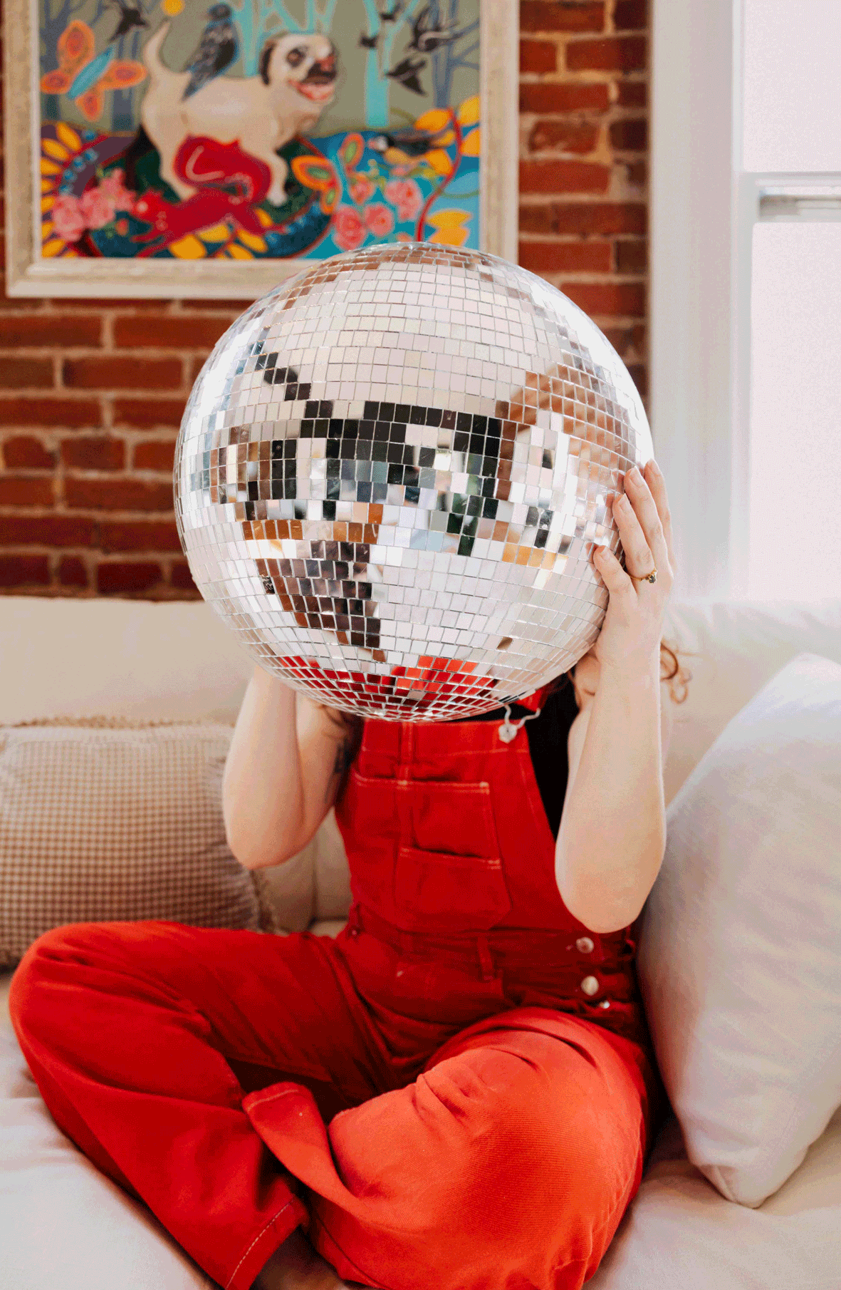 A woman sitting on a couch popping her head out from behind a disco ball.