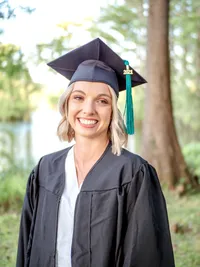 A smiling college senior in a graduation cap and gown standing by a lakeside in Wilmington, North Carolina. This portrait captures the joy of graduation, perfect for those looking for professional senior photography in a natural setting.