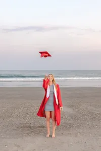 A college senior in a red gown joyfully tossing her graduation cap in the air on the beach with waves in the background in North Topsail Beach, North Carolina. This energetic image captures the celebration of graduation, perfect for memorable senior moments by the sea.