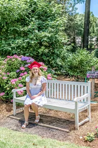 A high school senior in a red cap sitting on a white bench surrounded by blooming flowers in North Carolina. This charming scene captures the beauty of outdoor senior photography, perfect for those wanting vibrant and memorable graduation photos.