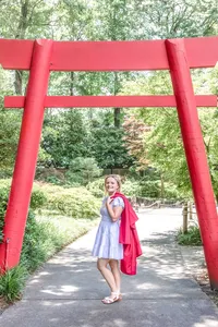 A high school senior holding her graduation gown over her shoulder, posing under a vibrant red Torii gate at the Arbortum in Wilmington, North Carolina. This unique setting highlights the charm of destination graduation photography, perfect for capturing distinctive senior photos.