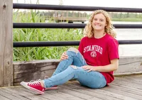 An Ashely High School senior wearing a Stanford Universtiy shirt sitting on a wooden dock with greenery in the background in Wilmington, North Carolina. This casual portrait reflects youthful spirit, perfect for those looking for relaxed and natural senior photos.
