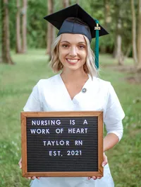 A UNCW nursing graduate in a cap and gown holding a message board that reads "Nursing is a Work of Heart" in a wooded area in Wilmington, North Carolina. This portrait captures academic achievement and personal dedication, perfect for celebrating graduation.