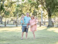 A family of four stands in a sunlit park near Wilmington, NC. The parents hold their young daughters, all dressed in pastel outfits, surrounded by lush greenery. This image highlights affordable family photography in Wilmington, NC, capturing warm family bonds.