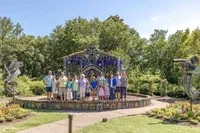 A family poses in front of a colorful mosaic fountain in a garden, surrounded by sculptures and greenery. Dressed in vibrant summer attire, they enjoy a sunny day, ideal for destination family photographer NC, capturing joyful and artistic moments.