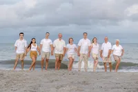 A large family stands hand in hand on the beach under a dramatic cloudy sky, dressed in white and khaki outfits. The ocean waves add a dynamic backdrop, perfect for North Carolina family photography, capturing unity and elegance.