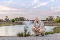 A college senior crouching by a lake with a fountain backdrop during a colorful sunset in Wilmington, NC. This relaxed portrait showcases professional senior photography, perfect for capturing natural and serene moments.