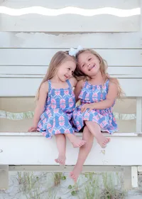 Two young sisters sit on a beach bench, wearing matching blue dresses with pink patterns and a white bow. Their joyful expressions capture a playful, sunny day by the sea, perfect for North Carolina family photography, highlighting sibling bonds.