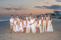 A large family dressed in white poses on the beach at sunset, with soft pastel skies and beachfront homes in the background. This image captures the elegance and warmth of a family gathering, perfect for artistic family photography in Wilmington, NC.