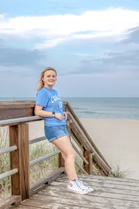 A high school senior in a carolina blue UNC shirt and shorts leaning on a boardwalk railing with the ocean in the background in Carolina Beach, North Carolina. This casual and vibrant portrait captures the relaxed vibe of senior photography, perfect for beach-themed sessions.